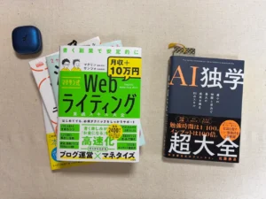 テーブルに置かれたワイヤレスイヤホンと、積まれたWebライティングの本、そして今日読む1冊として出された『AI独学超大全』。50代の自動化学習の環境。