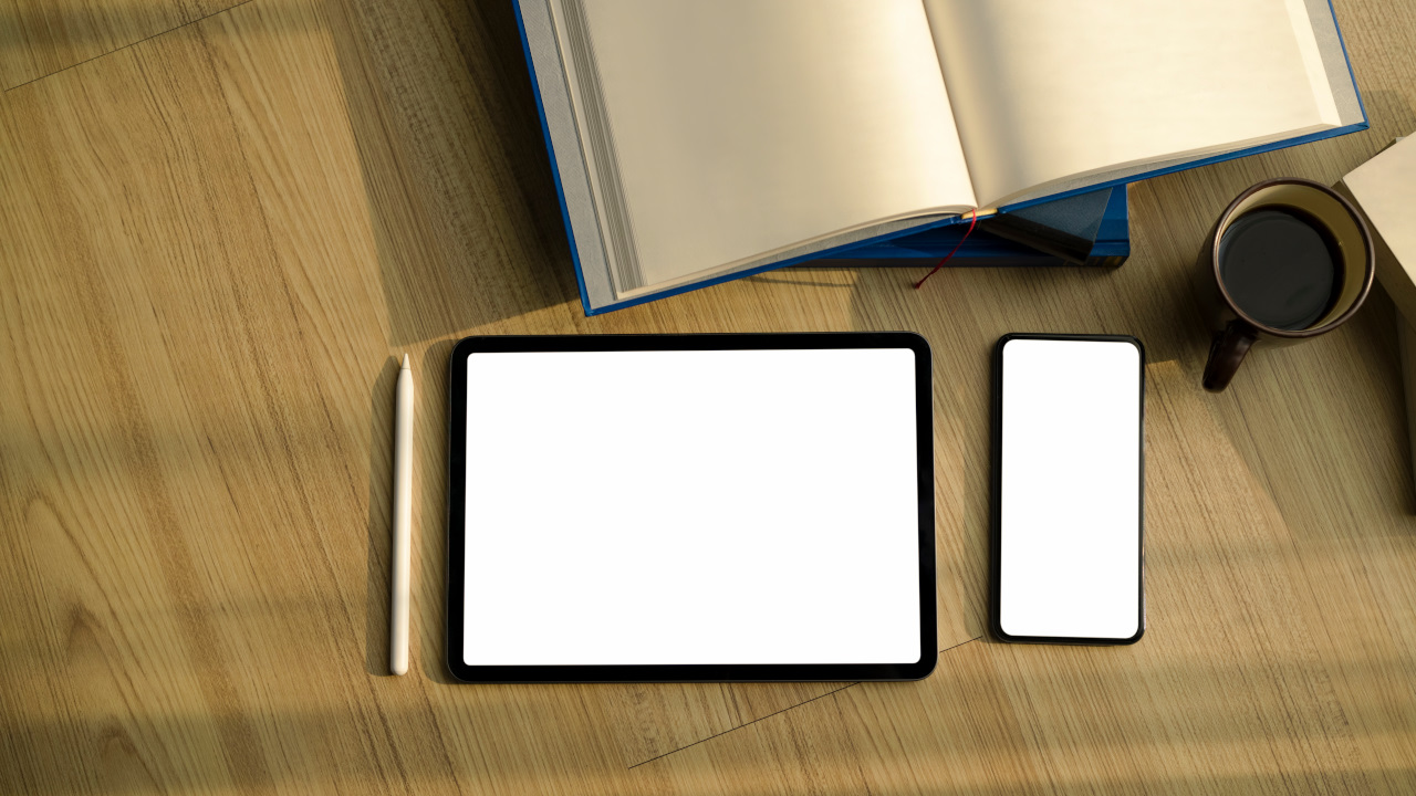 木漏れ日が差し込む木製のデスクの上に置かれた、開いた紙の本とタブレット端末、スマートフォン